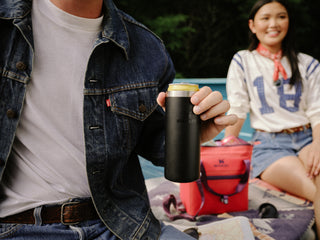 Close-up of a person holding a black Stanley can cooler with a drink, sitting on a picnic blanket with another person and a red Stanley cooler in the background.