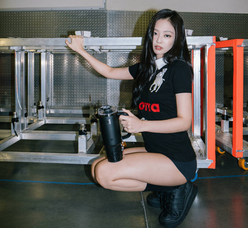 Jennie of BlackPink in a black t-shirt and boots holding a black Stanley tumbler while crouching in an industrial setting.